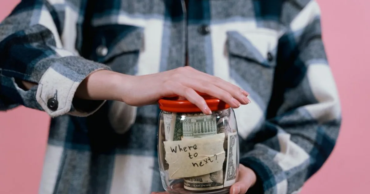 Close-up of a woman holding a savings jar labeled 'Where to next?' filled with US dollar bills.