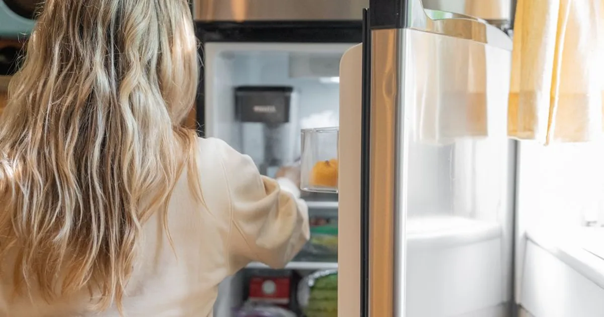 Back view of a blonde woman retrieving food from a refrigerator in a bright kitchen.