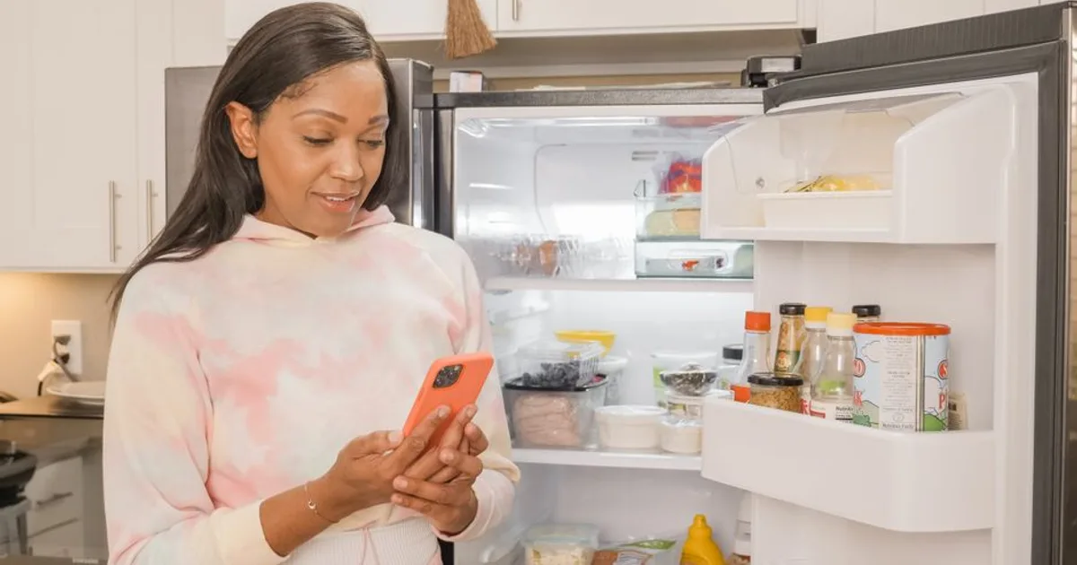 Woman using smartphone for online shopping while standing in the kitchen with an open refrigerator.