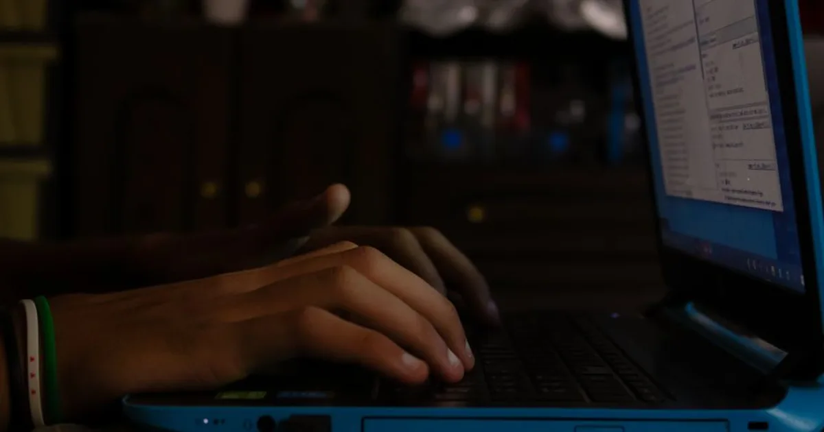 Hands typing on a blue laptop indoors, showcasing focus on work or study in a dimly lit room.