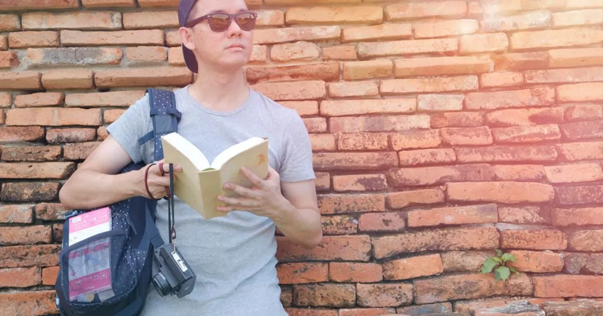 A traveler holding a book stands by an ancient brick wall in sunlight, capturing a moment of exploration.